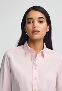 Camicia rosa chiaro con design a bottoni, colletto a punta e maniche lunghe. Presenta un piccolo dettaglio ricamato sul lato sinistro del petto.