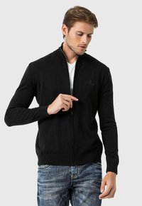 Schwarze Reißverschlussstrickjacke mit geripptem Kragen, langen Ärmeln und dezentem Logo auf der Brust. Kombiniert mit blauen Jeans, zeigt einen lässigen Stil.
