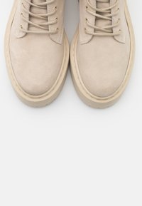 Beige semišové kotníkové boty skulatými špičkami, s odpovídajícími tkaničkami a silnou krémovou gumovou podrážkou. Hladký povrch s minimem švů.