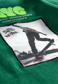 Grüne Sweatshirt-Textilie mit einem rechteckigen schwarzen Patch, auf dem ein Graustufen-Bild eines Skateboards und Text im Graffiti-Stil am unteren Rand zu sehen ist.