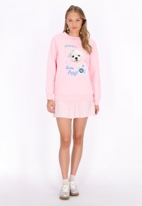 Világos rózsaszín pulóver kölyökkutyás grafikával és "Aranyos Kölyökkutya" felirattal, világos pliszírozott szoknyával és ezüst tornacipővel párosítva.