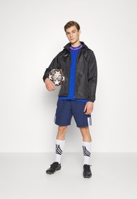 Black lightweight jacket, blue shirt, navy shorts with white stripes, black soccer cleats, white socks with black pattern, holding soccer ball.