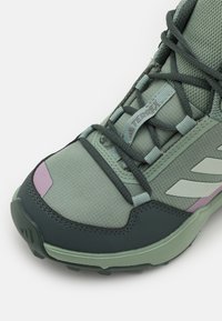 Zaļi sporta apavi ar teksturētu sietiņu augšdaļu, pastiprinātu purngalu un purpura akcentu. Iekļauta gaiši zaļa zole un melni saķeres uzgaļi.
