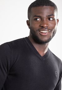 Schwarzer, figurbetonter V-Ausschnitt-Pullover aus einem glatten Strickstoff, mit einem dezenten Logo auf der Brust und gerippten Details am Ausschnitt.