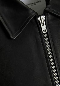 Black leather jacket featuring a pointed collar and a silver metal YKK zipper. The texture is smooth with a matte finish.