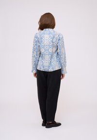 Camicia a maniche lunghe blu con motivo, che presenta disegni intricati in beige. Indossata con pantaloni neri larghi e scarpe marroni.