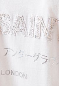 Biała bawełniana koszulka z teksturowanym przeszyciem, z haftowanym konturem napisu "SAINT", srebrnym nadrukiem z tekstem, w tym japońskimi znakami oraz "LONDON".