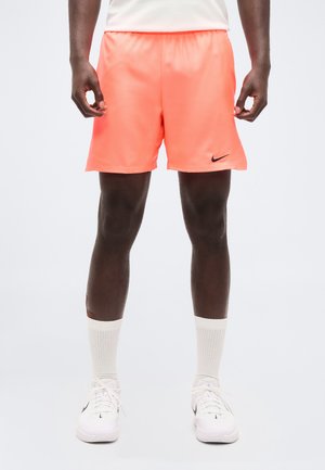 Męski model w jasnych koralowych szortach Nike, białe skarpetki za kostkę, białe sneakersy Nike, stoi na jednolitym jasnym tle.