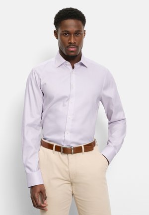 Homme portant une chemise boutonnée violet clair rentrée dans un pantalon beige avec une ceinture en cuir marron, debout devant un fond blanc.