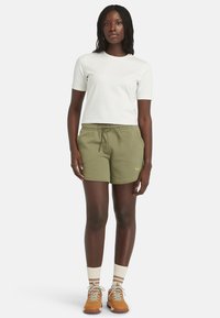 Camiseta de algodón en blanco, combinada con pantalones cortos verde olivo que cuentan con una cintura elástica. Lleva zapatos naranjas y calcetas a rayas en color crema.