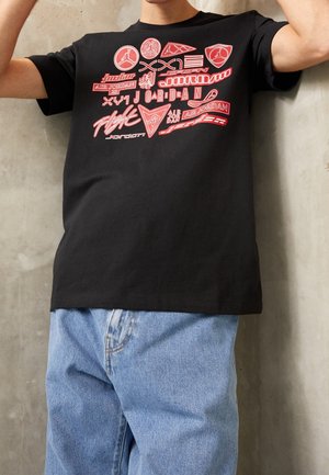 Person wearing a black t-shirt with multiple red and white Air Jordan logos and texts, paired with light blue jeans against a gray wall.
