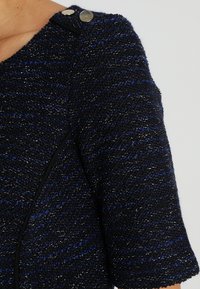 Marineblå og sort textured jakke med metaltråde, med en rund halsudskæring og to metalliske knapper på skulderen.