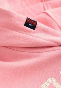 Un tissu rose doux avec une étiquette de marque "Superdry" noire et rouge cousue près d'un bord plié et un motif texturé partiel en blanc.