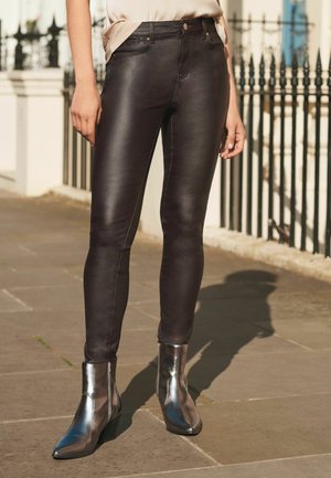 Crne uske traperice s sjajnom završnicom, sa prednjim džepovima i metalnim akcentima, u kombinaciji s metalnim čizmama do gležnja.