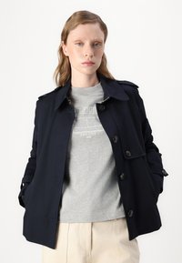 Navy trench coat with a classic cut, double-breasted buttons, and shoulder epaulets, worn over a grey graphic t-shirt and off-white trousers.