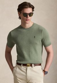 Homme portant un pull à manches courtes côtelé vert, pantalon beige, ceinture marron, montre et lunettes de soleil, debout les mains dans les poches.