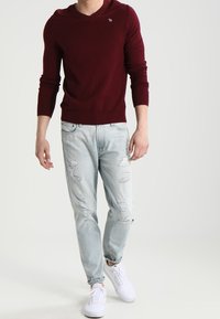 Burgundy stickad tröja med v-ringning och liten logga, i kombination med ljusblå slitna jeans och vita sneakers. Avslappnad outfit.