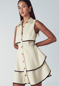 Abito senza maniche beige con gonna a strati e bordi neri, con bottoni frontali, indossato da una donna con lunghi capelli scuri e orecchini a cerchio dorati.