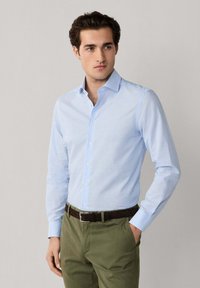 Homme portant une chemise à rayures bleu clair à boutons et un pantalon vert olive avec une ceinture foncée, debout avec une main dans la poche.