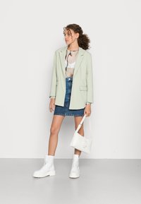 Blazer verde menta sobre uma blusa de padrão claro, combinado com uma saia de ganga, botas de combate brancas e uma pequena mala branca.