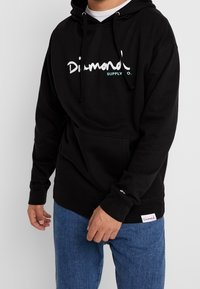 Muž oblečený v černé mikině Diamond Supply Co. s přední kapsou a modrými džíny, stojící před jednoduchým bílým pozadím.