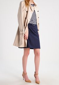 Impermeable beige sur une jupe bleu marine et un haut rayé. Talons hauts marron clair avec des brides autour de la cheville. Textures lisses dans l'ensemble.