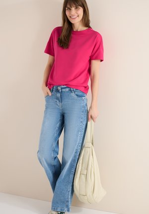 Femme souriante, portant un t-shirt rose vif et un jean bleu, tenant un sac à main beige clair sur un fond uni beige.