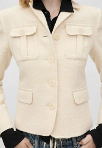 Chaqueta de tweed crema con un corte estructurado, que cuenta con dos bolsillos frontales, cuatro botones y un collar con muesca, combinada con una camisa negra debajo.