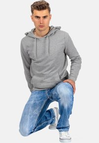 Grijze katoenen hoodie met een trekkoordkap, gecombineerd met vervaagde blauwe spijkerbroek en witte sneakers, in een knielende pose tegen een witte achtergrond.