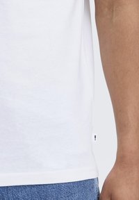 White cotton t-shirt with short sleeves and a straight hem. Features a small branded tag on the side. Worn with blue denim jeans.