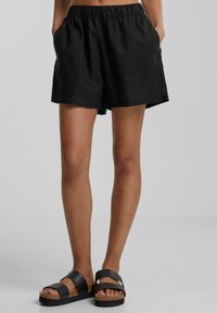 Personne portant un short noir à taille élastique avec les mains dans les poches et des sandales à plateforme noires devant un fond uni.