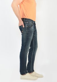 Vaqueros de mezclilla en azul oscuro con un corte ajustado, que presentan secciones desgastadas, bolsillos delanteros y costuras en naranja contrastante. Combinados con zapatillas blancas.