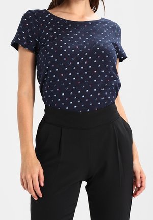 Kobieta w granatowej bluzce z krótkim rękawem ozdobionej małymi różowymi i białymi serduszkami, wpuszczonej w wysokie, czarne spodnie.