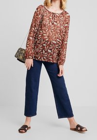Blouse marron à manches longues et transparente avec des motifs de feuilles, associée à un jean large bleu foncé et des sandales à glisser marron. Sac à main à motifs noir et or.