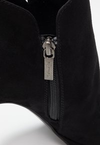 Schwarze Wildleder-Stiefelette mit einem seitlichen Reißverschluss, die eine schlanke Form, minimale Nähte und eine glatte Textur aufweist. Der Reißverschluss-Anhänger hat ein Branding-Detail.