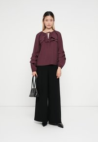 Bordeaux gekruide blouse met lange mouwen, ronde hals en strikdetail, gecombineerd met zwarte wijde broek en een kleine zwarte handtas.