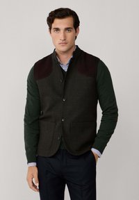 Gilet verde scuro con tessuto testurizzato, toppe in pelle marrone sulle spalle e bottoni. Indossato sopra un maglione verde e una camicia a righe.