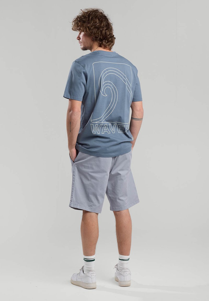 Light blue cotton t-shirt with white wave graphic and "WAVES" text on back, paired with gray shorts and white sneakers with green accents.