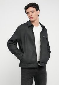 Μαύρο μπουφάν με φερμουάρ και γιακά, με τσέπη στο στήθος και πλαϊνές τσέπες, κατασκευασμένο από λείο ύφασμα, συνδυασμένο με λευκό μπλουζάκι και σκούρο τζιν.