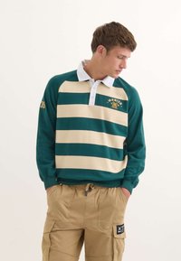 Zeleno-beige pruhovaná dlouhá polo košile s bílým límečkem a vyšíváním 'Mnichov 1939'. Látka vypadá měkce, má žebrované manžety.