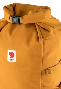 Oranssi rullaläppäreppu, joka on valmistettu teksturoidusta kankaasta. Varustettu mustalla muovisoljella ja logopatchilla. Sisältää sivuhihnoja säätämistä varten.