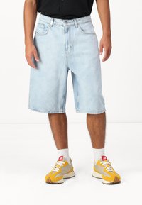 Pantalones cortos de mezclilla azul claro con un corte holgado, que cuentan con un cierre frontal, diseño de cinco bolsillos y un dobladillo recto. Llevado con zapatillas amarillas y grises.