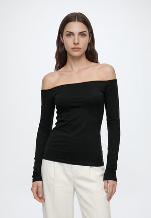 Femme aux longs cheveux bruns porte un top noir à épaules dénudées et manches longues et un pantalon blanc, debout sur un fond clair uni.