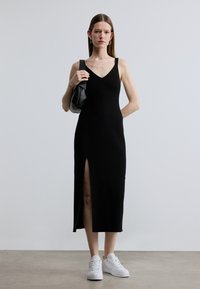 Čierne rebrované šaty bez rukávov s V výstrihom, s bočným rozparkom, nosené s bielymi teniskami a doplnené čiernou taškou.
