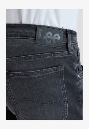 Nærbilde av svarte denimjeans bakside med Lee-merke i skinn og liten tekstiletikett, brukt med en innstøpt hvit skjorte.