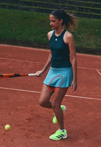 Tennis-Outfit mit einem dunklen Tanktop und einem hellblauen, plissierten Rock. Helle grüne Sportschuhe mit dunklen Streifen und ein Tennisschläger.