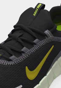 Κοντινή λήψη μαύρου αθλητικού παπουτσιού Nike Free με λογότυπο Swoosh σε νέον κίτρινο και μαύρα κορδόνια σε λευκή σόλα.