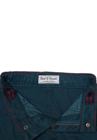 Teal puuvillahousut punaisella tikkauksella, joissa on nappi, vetoketjulento ja sisätasku. Täggissä lukee "Band of Rascals."