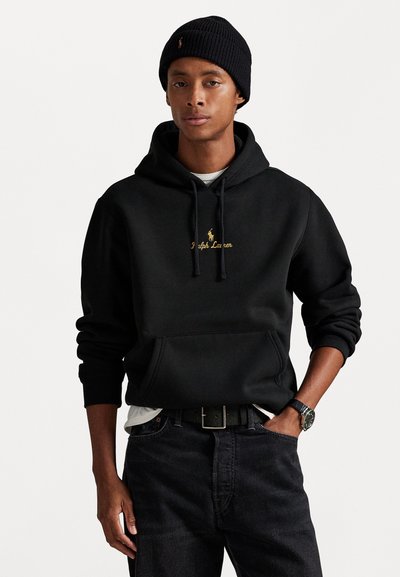 Sort hættetrøje med en frontlomme og snørebånd i hætten, med gyldent tekst "Ralph Lauren" og et logo, båret over en grå skjorte og mørke jeans.