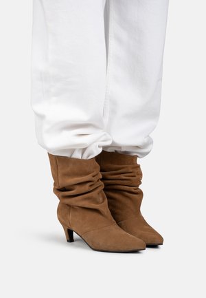 Botines marrones de ante con tacón medio y un estilo holgado, combinados con pantalones blancos y sueltos sobre un fondo claro.
